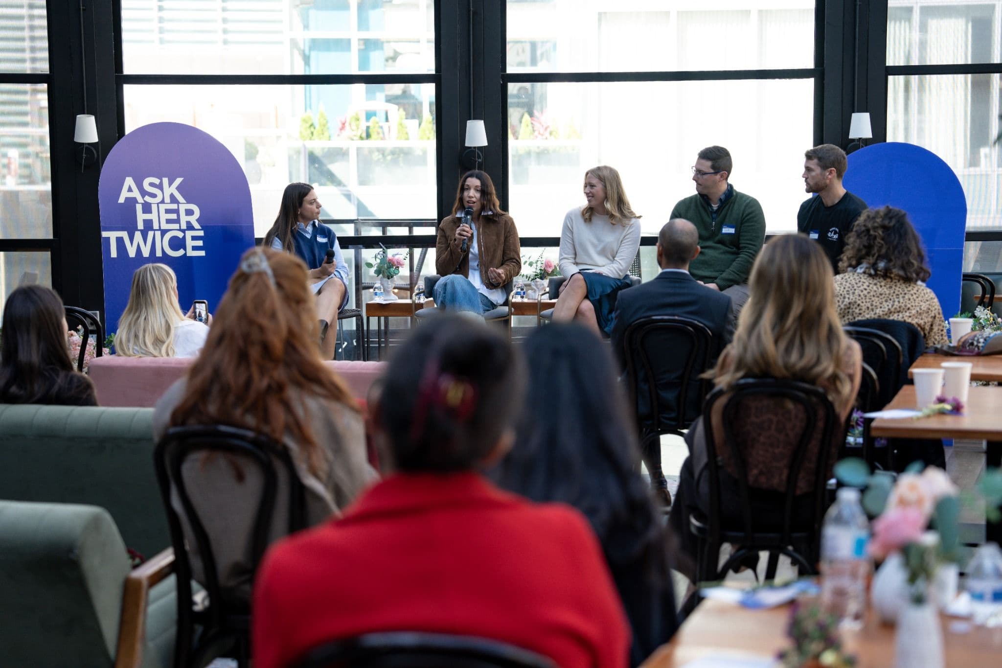 Guests seated at tables face a five-person panel in front of a large Ask Her Twice banner