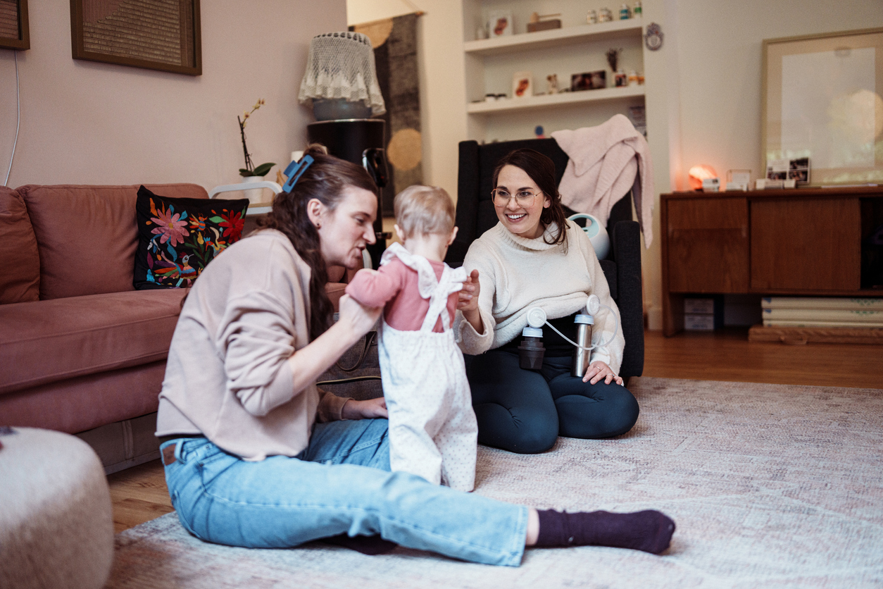 A woman sitting on the floor visiting a friend who is holding her toddler, with a breast pump nearby