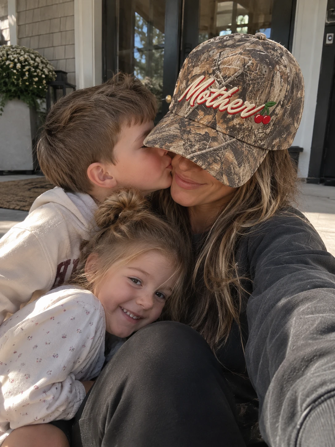 A woman in the Mother Hat smiling as a young boy kisses her cheek and a little girl leans on her lap