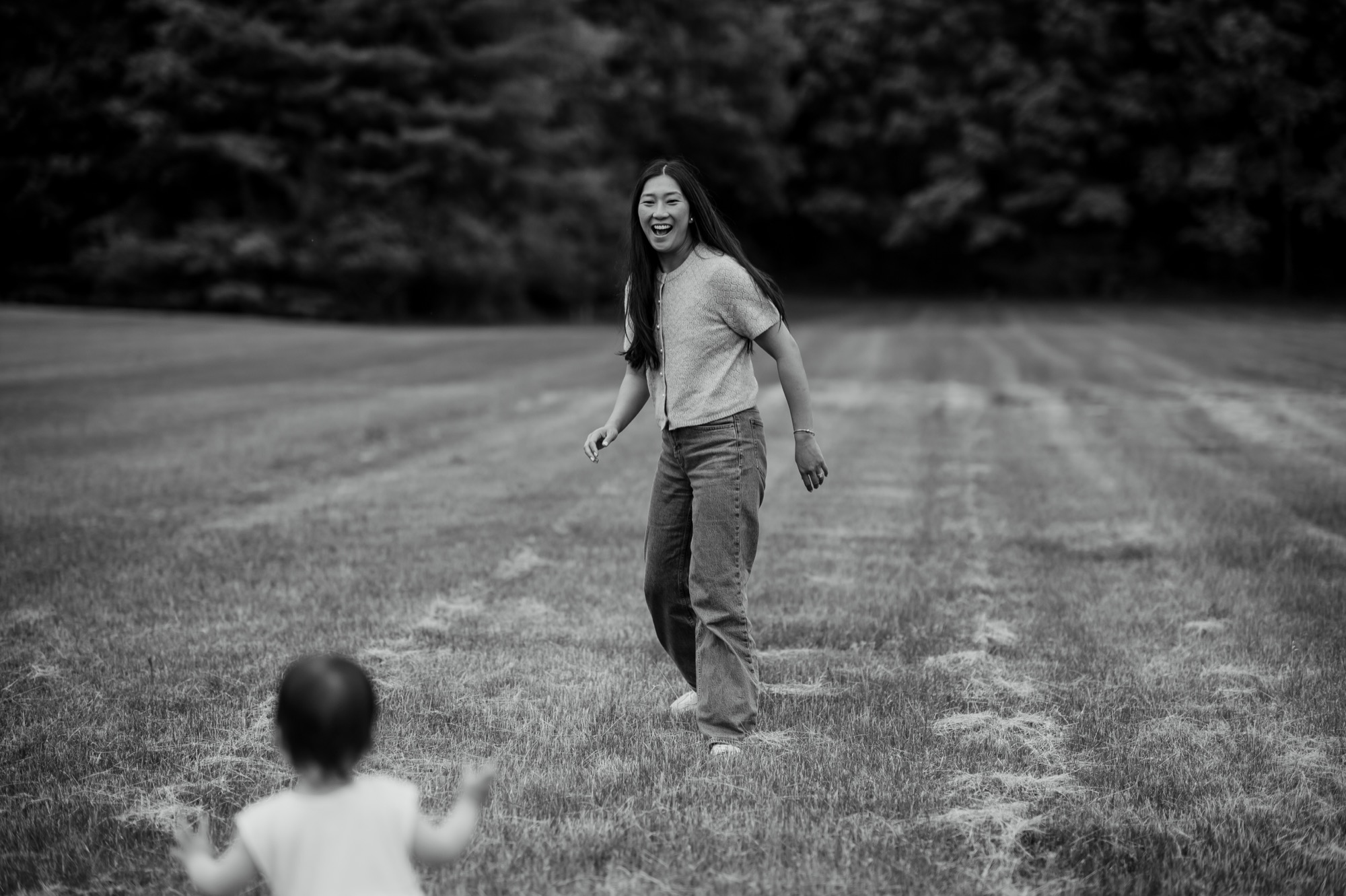 A mother laughing in a field as her toddler walks toward her.